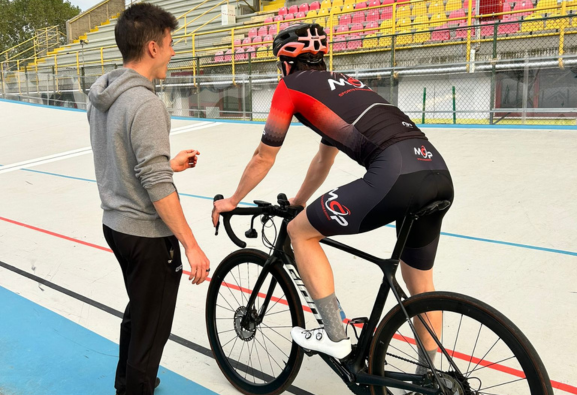 Un ciclista su una bici da pista parla con un allenatore vicino al bordo della pista di un velodromo.